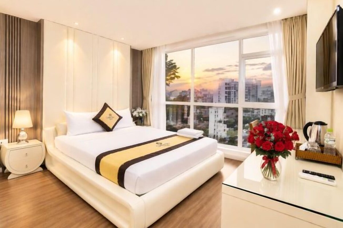 A hotel room with a bed, a nightstand, a desk, a TV, and a window with a city view.