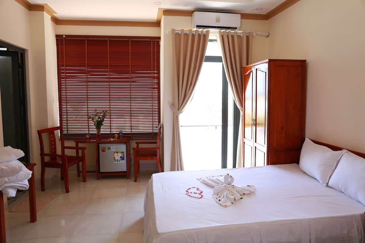 A hotel room with a bed, a desk, a chair, a small refrigerator, and a window with blinds.