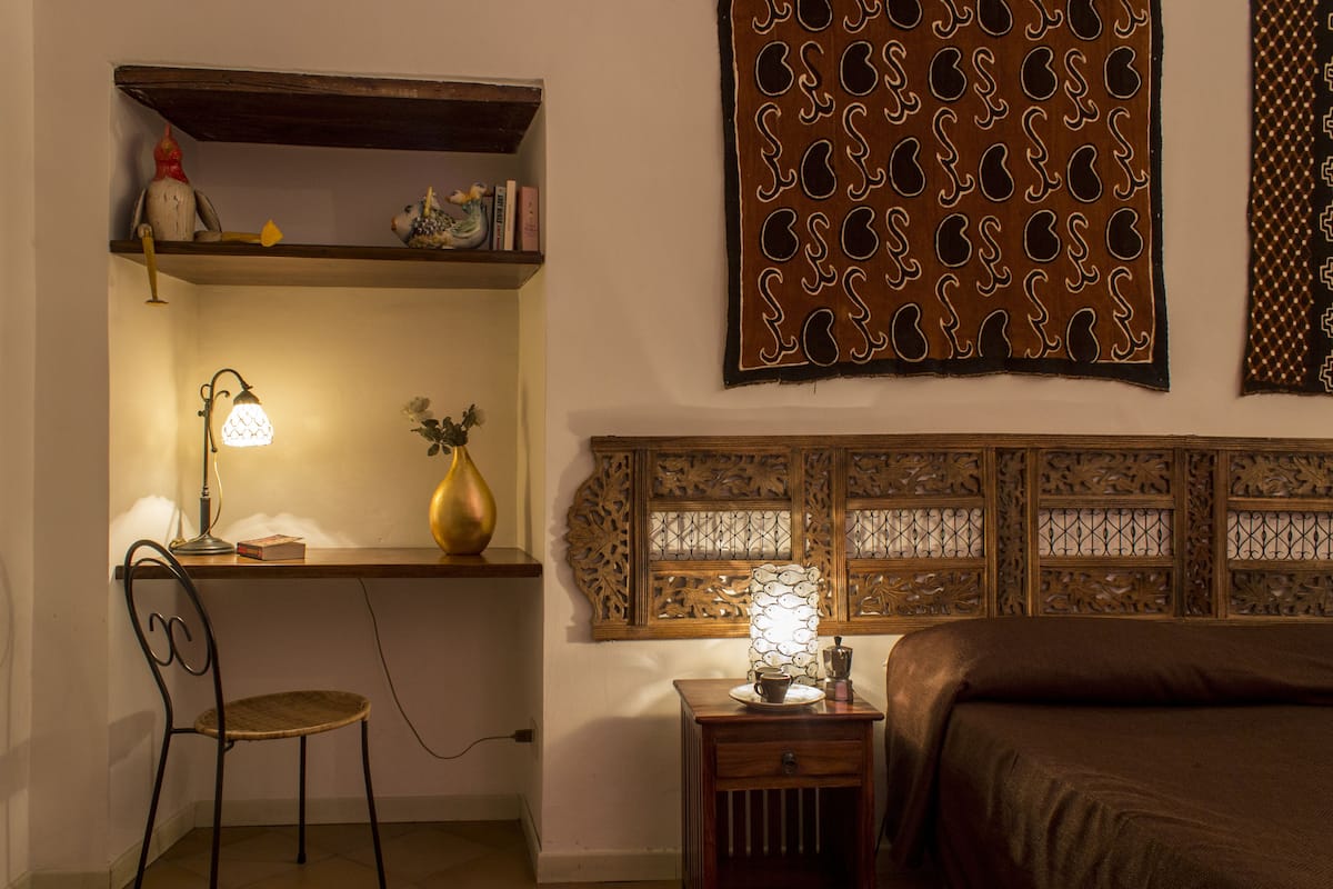 A bedroom with a bed, a desk with a lamp, a chair, a shelf with decorative items, and a patterned wall hanging.