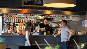 A bar area with a bartender serving drinks, a menu board above the bar, and various bottles and glasses on the shelves.