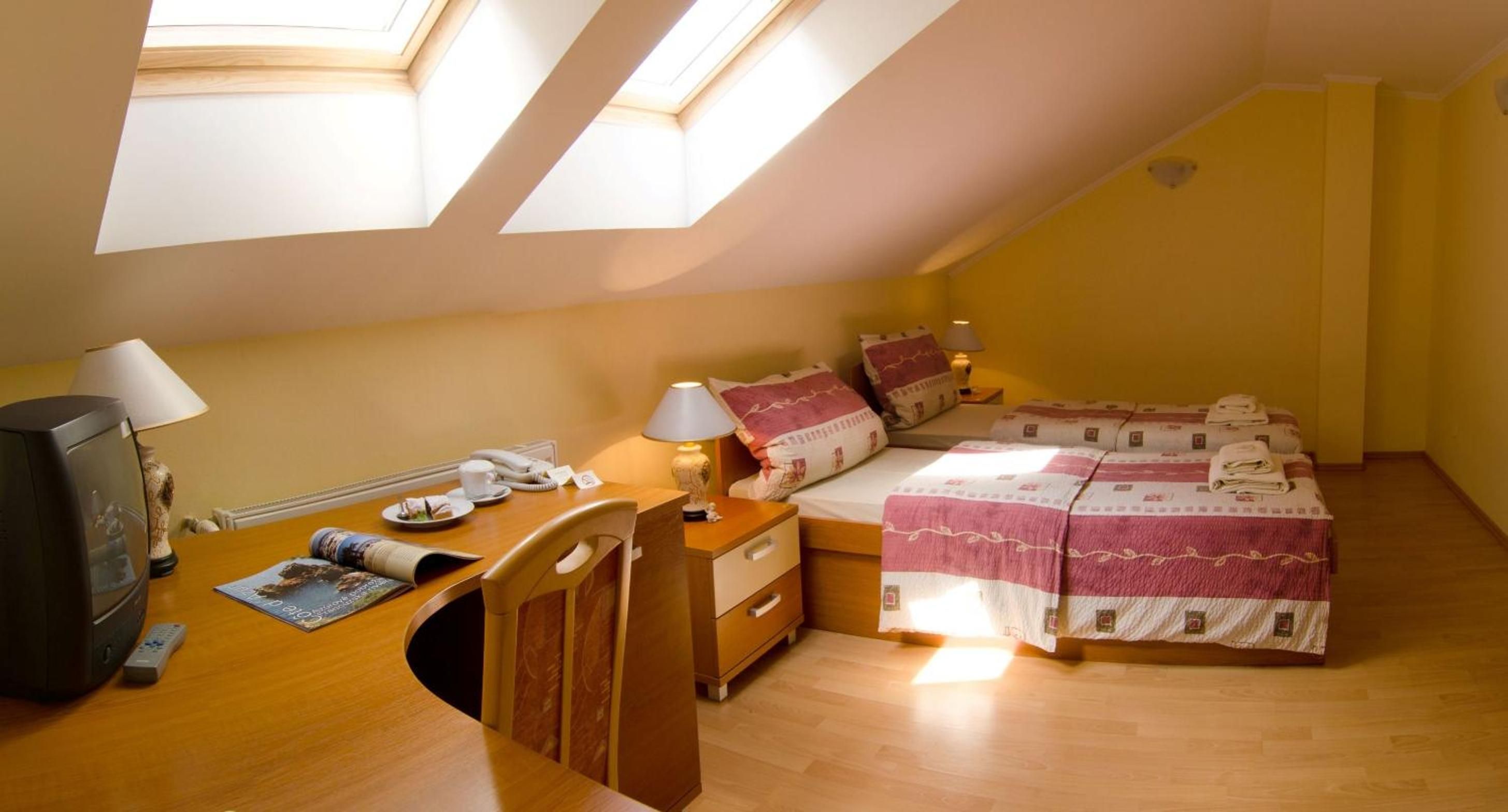 A room with a bed, bedside table, lamp, and a desk with a computer and remote control.