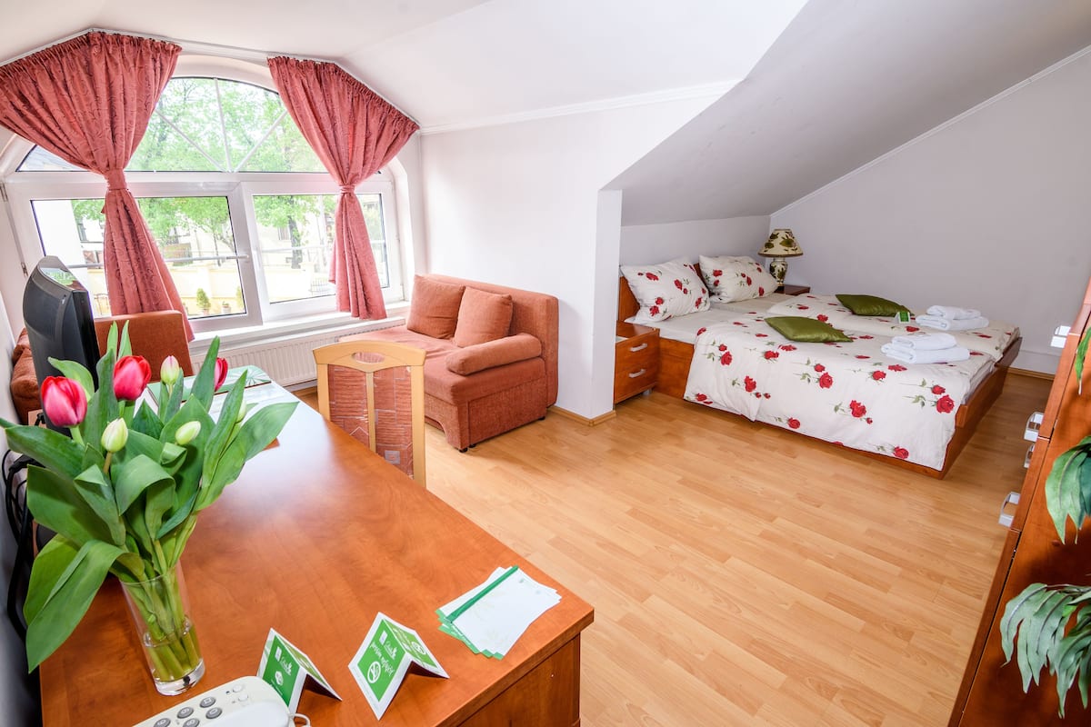 Ein Schlafzimmer mit Holzboden, einem Bett mit floralem Bettzeug, einem Schreibtisch mit Blumenvase und einem Fenster mit Vorhängen.