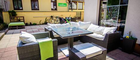Un patio al aire libre con mobiliario de mimbre, una mesa de vidrio y vistas a un edificio amarillo con un letrero que dice "DEPENDANCE".