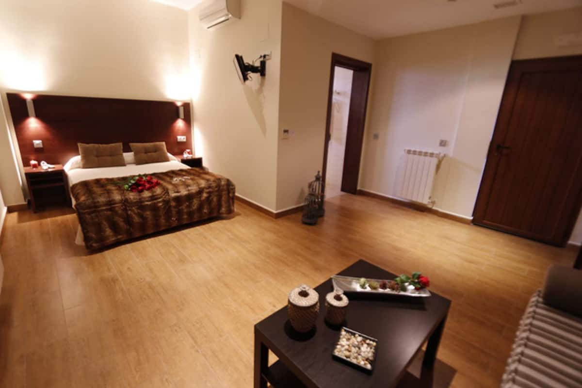 Une chambre avec un plancher en bois, un lit avec une tête de lit marron, des tables de chevet avec des lampes, une table de nuit, une commode, une table en bois ornée d’objets décoratifs et une porte donnant sur une autre pièce.