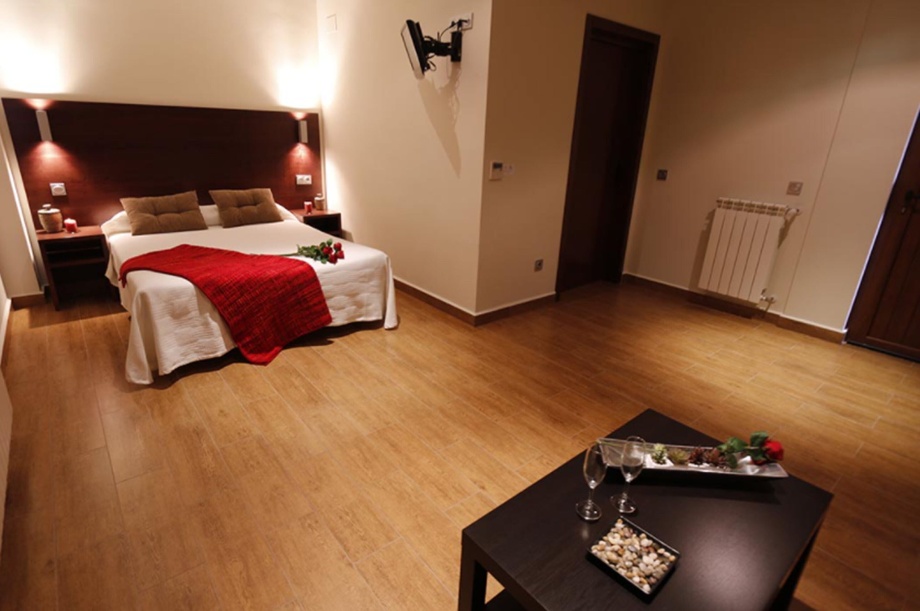 Habitación de hotel con una cama grande, una mesita de noche con lámpara, una pequeña mesa con una copa de vino y aperitivos, y un radiador en la pared.