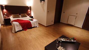 Habitación de hotel con una cama grande, una mesita de noche con lámpara, una pequeña mesa con una copa de vino y aperitivos, y un radiador en la pared.