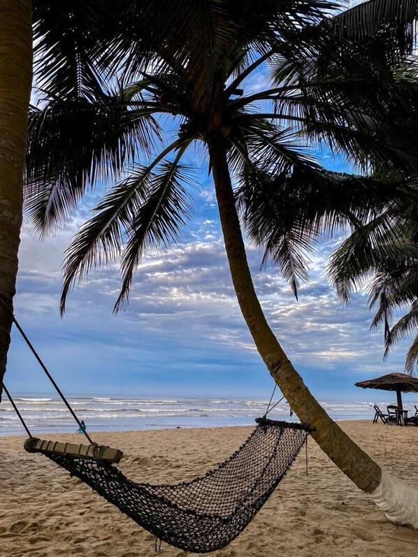 Una hamaca atada entre palmeras sobre una playa arenosa, con cielo despejado.