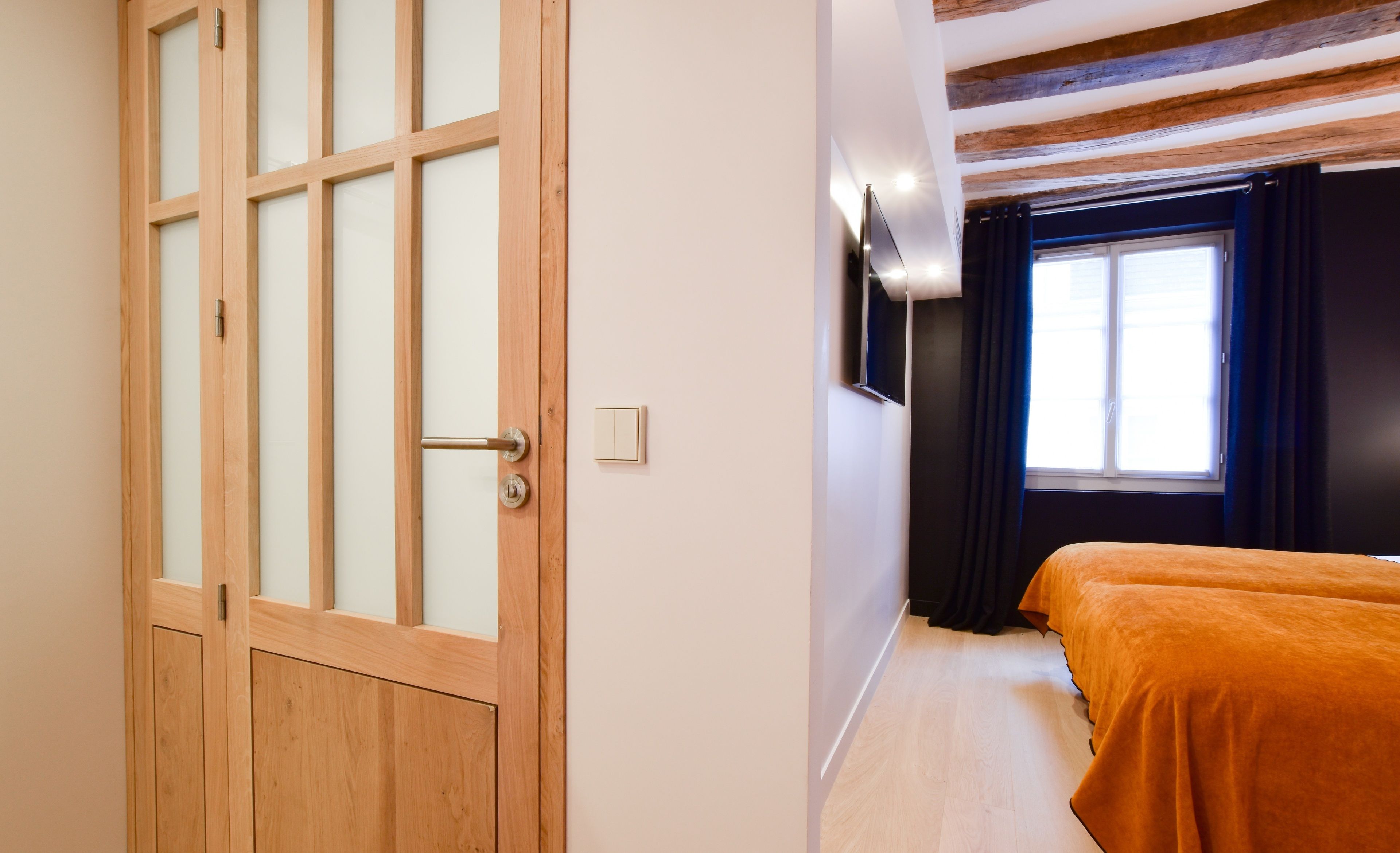 A room with a wooden door, a bed with an orange cover, a window with curtains, and a wall-mounted light fixture.