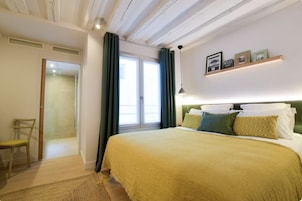 Un dormitorio con una cama, una silla, un ventanal con cortinas y una estantería con fotografías.