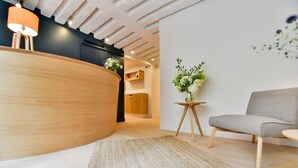 Et moderne receptionområde med en træreception, en grå sofa, et lille rundt bord med en vase blomster og en struktureret måtte på gulvet.
