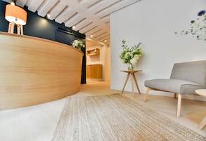 Una moderna zona de recepción con un mostrador de madera, un sofá gris, una pequeña mesa redonda con un jarrón de flores y una alfombra texturizada en el suelo.