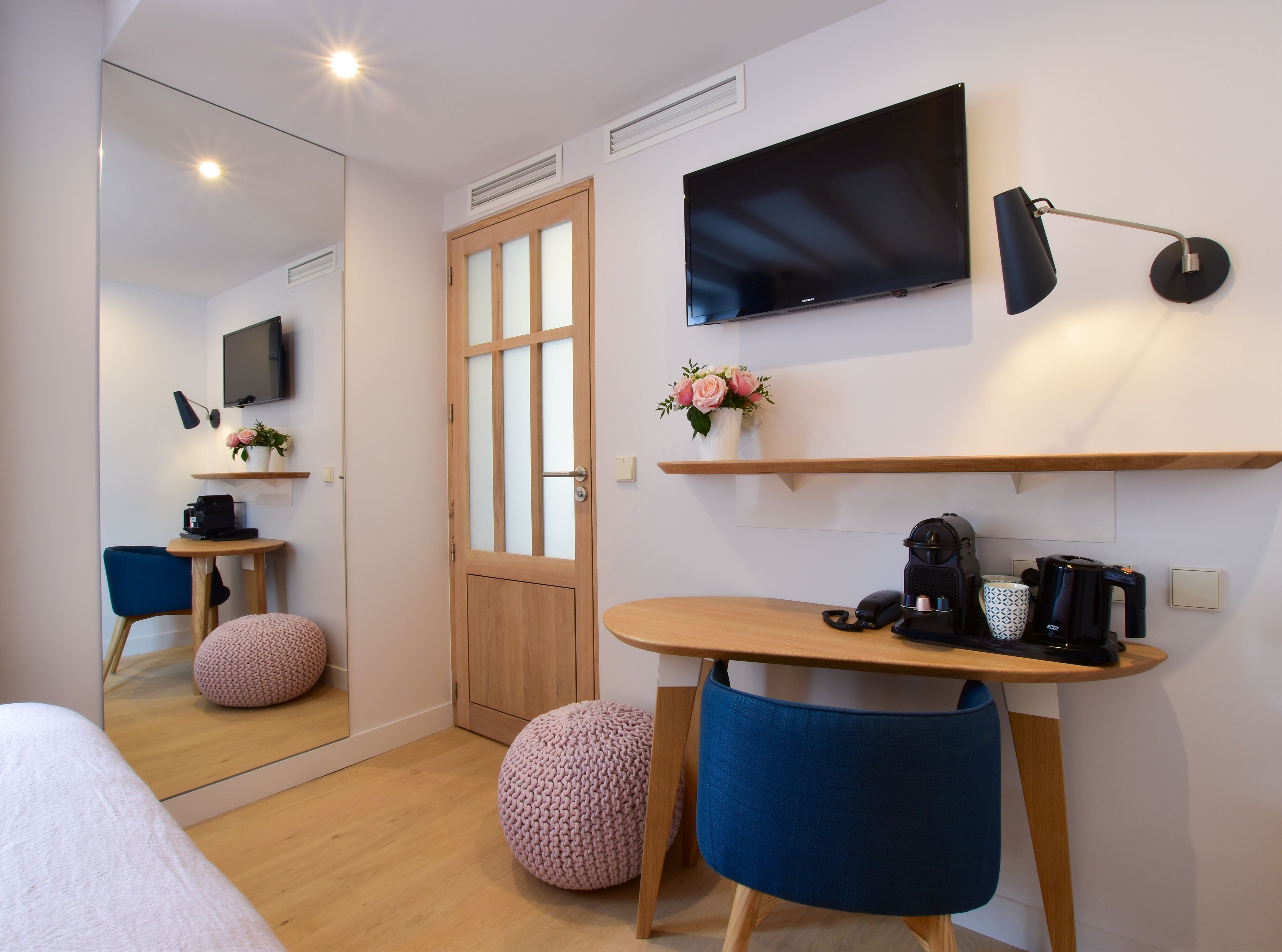 Una habitación de hotel moderna con un espejo grande, un televisor de pantalla plana, un escritorio de madera, una silla azul, un otoman rosa y una cafetera.