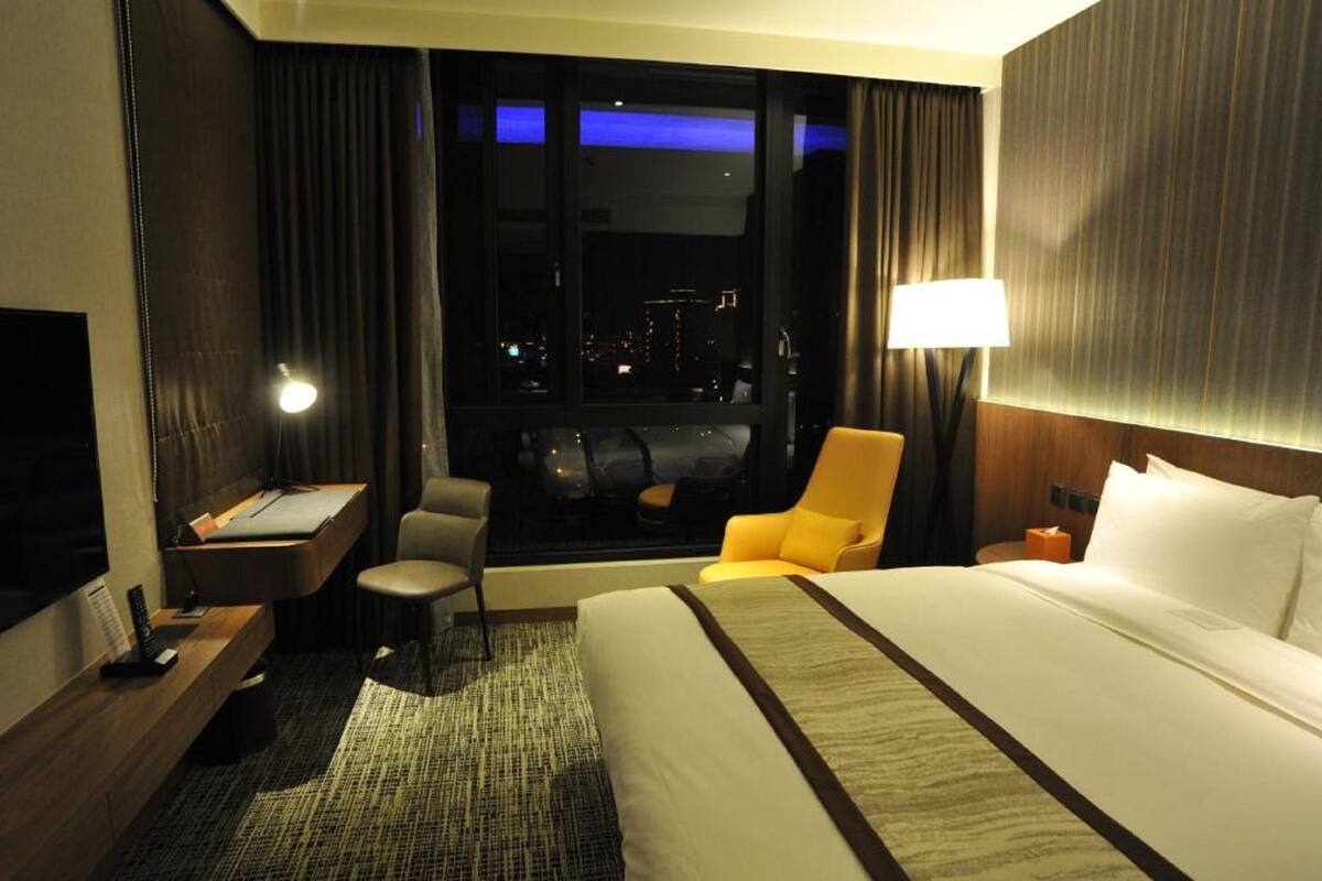 A hotel room with a large bed, a desk with a chair, a small table, and a window with a city view.