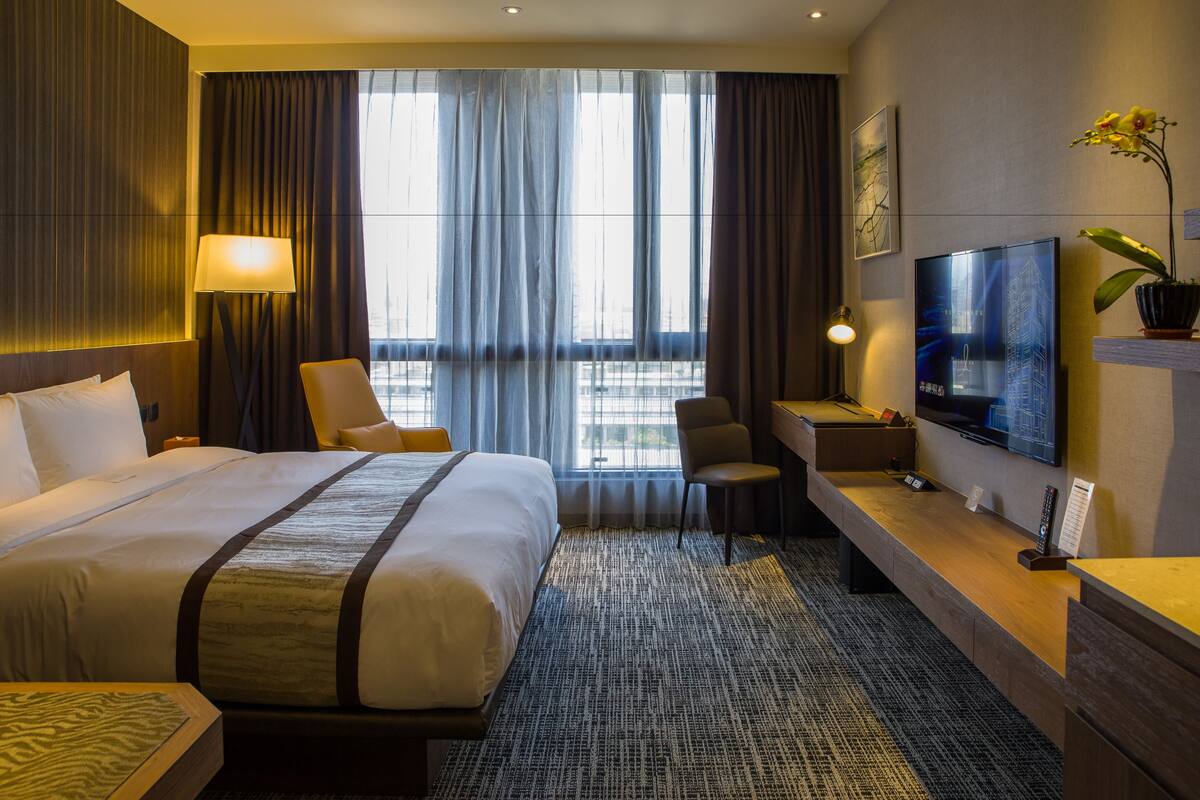 A hotel room with a large bed, a desk, a chair, a TV, and a window with curtains.