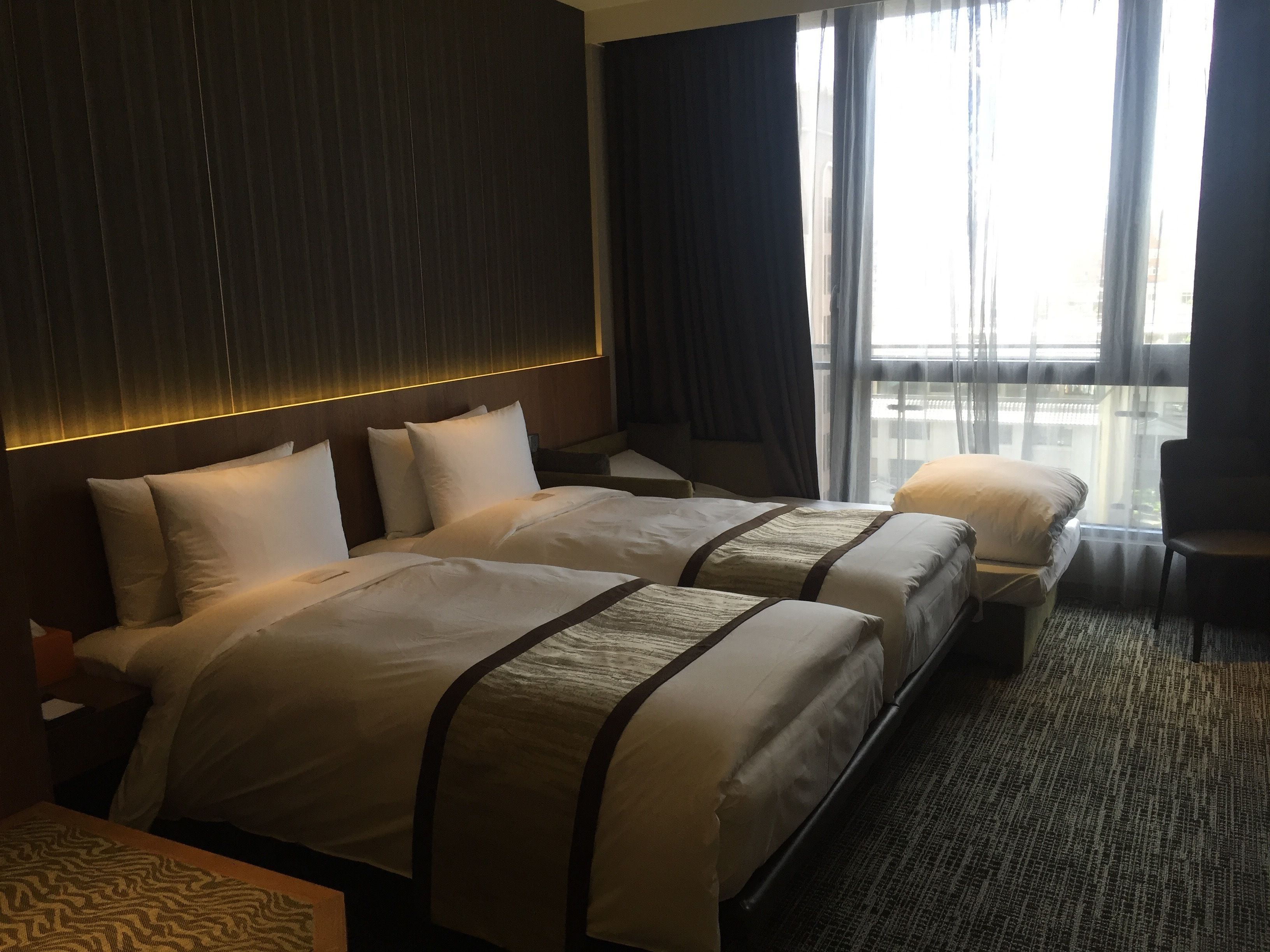 A hotel room with two beds, a wooden headboard, a large window with curtains, and a carpeted floor.