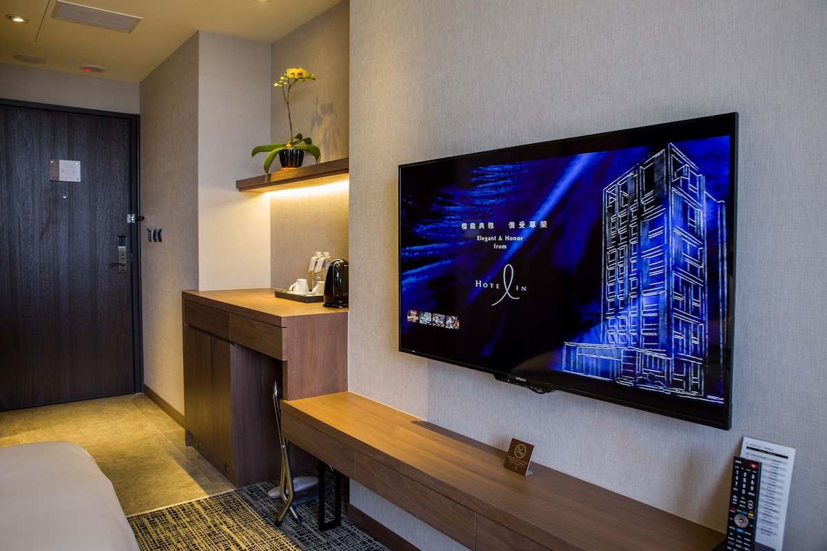 Habitación de hotel con cama, un banco de madera, un televisor montado en la pared mostrando un edificio y una pequeña planta en maceta sobre una estantería.