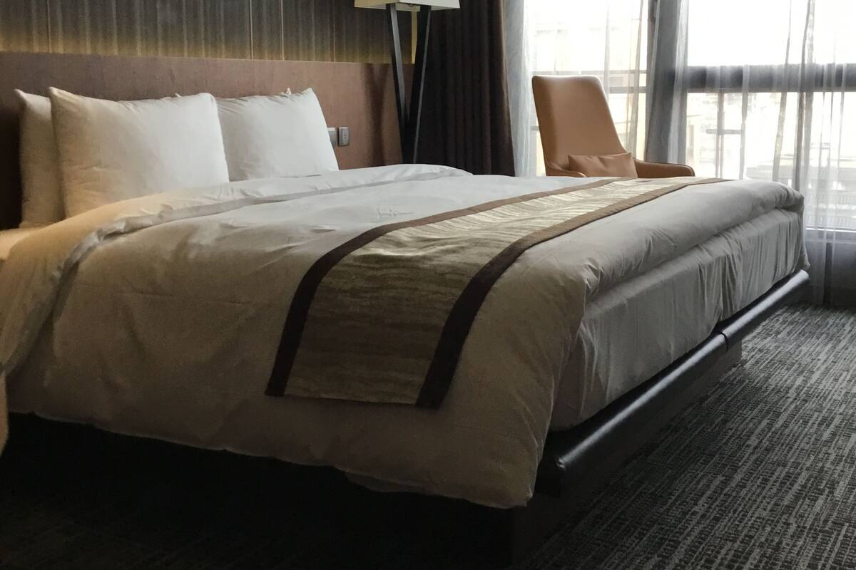 A hotel room with a large bed, a standing lamp, a chair, and a view of the city through large windows.