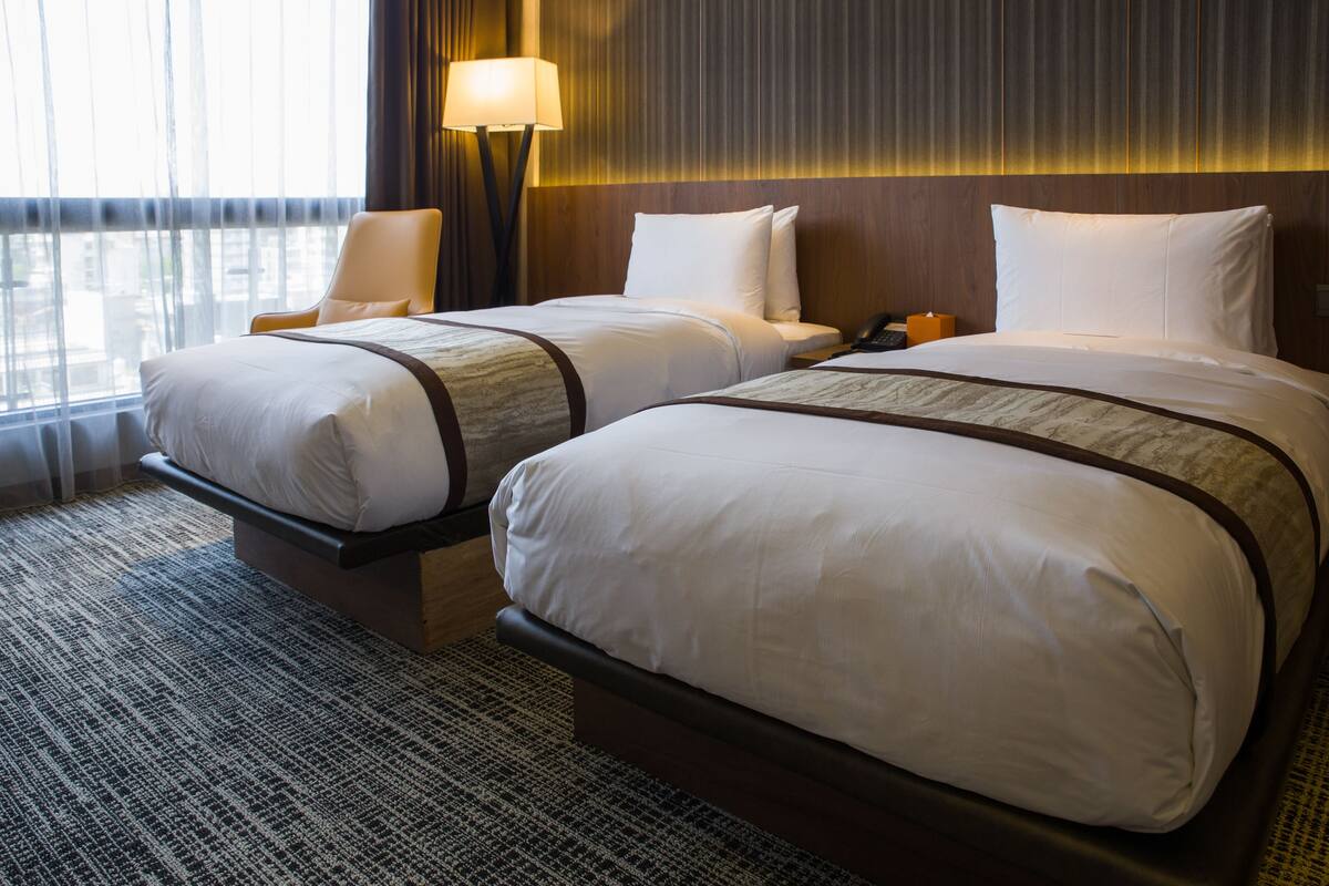 A hotel room with two beds, a nightstand, a lamp, and a view of the city through large windows.