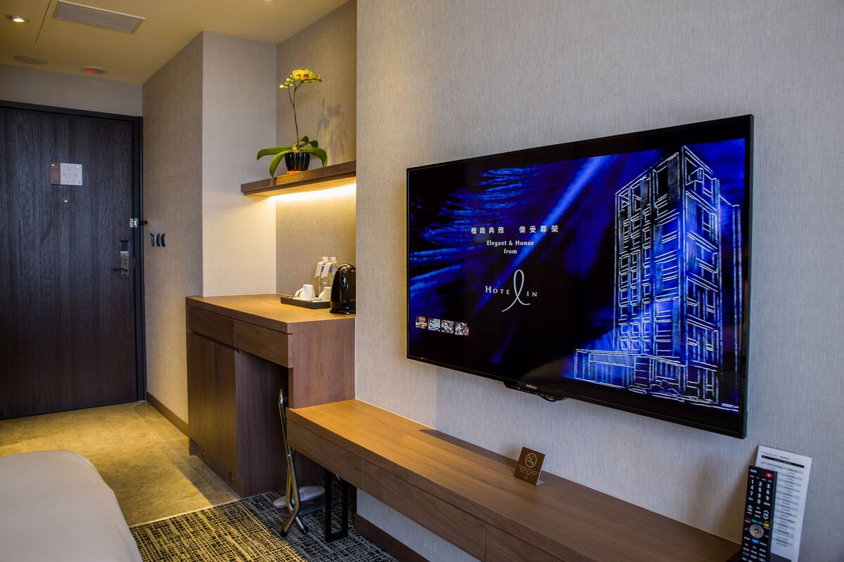 A hotel room with a large flat-screen TV mounted on the wall displaying a building image, a wooden bench, a small table with a plant, and a door with a lock.