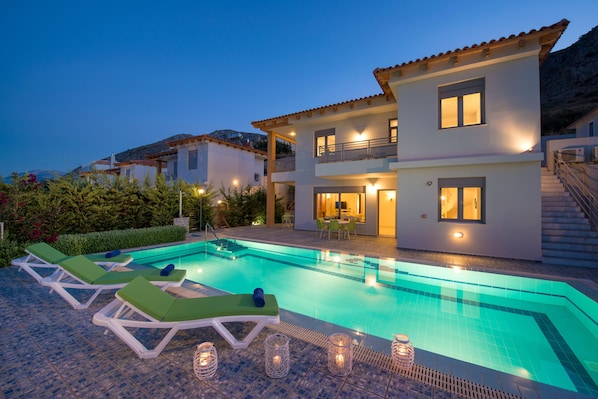 A modern two-story house with a swimming pool, lounge chairs, and a well-lit patio area. - Danae's Luxury Villas (Hersonissos)