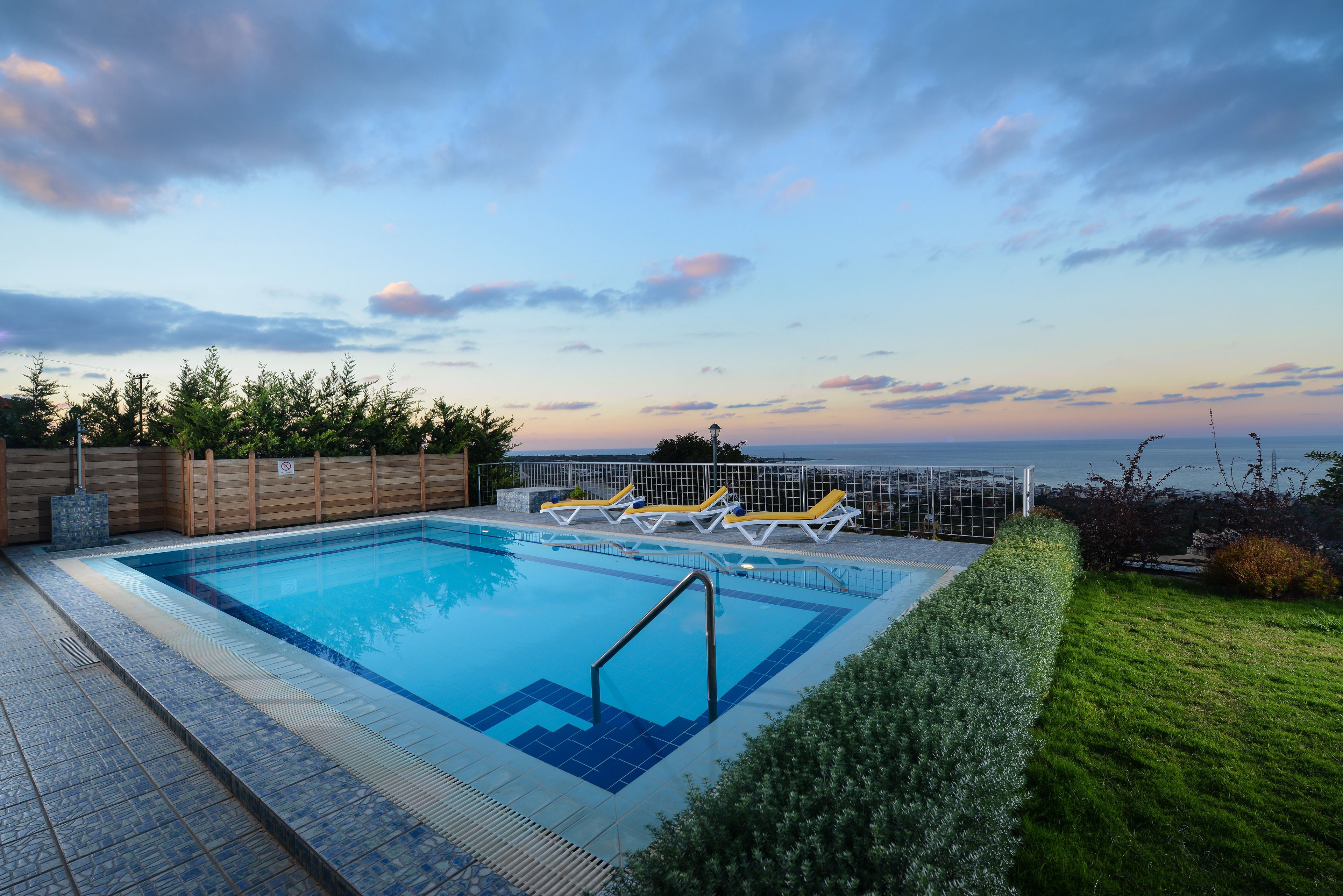 A rectangular swimming pool with a blue tiled edge, surrounded by a wooden fence and a metal railing. There are three sun loungers with yellow cushions and white frames. The pool is adjacent to a well-maintained grassy area with a hedge.