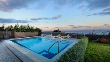 A rectangular swimming pool with a blue tiled edge, surrounded by a wooden fence and a metal railing. There are three sun loungers with yellow cushions and white frames. The pool is adjacent to a well-maintained grassy area with a hedge.