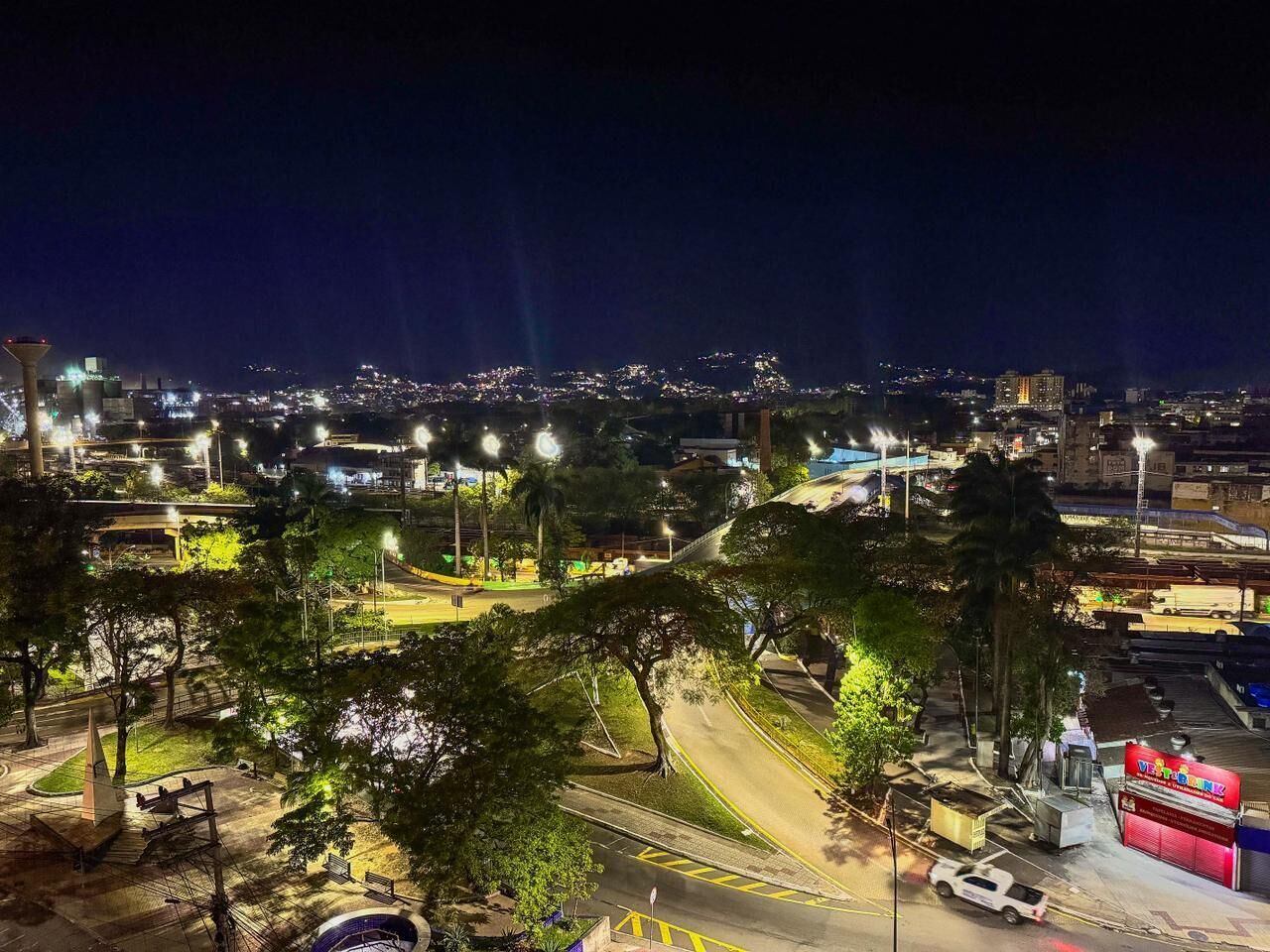 A nighttime cityscape with illuminated buildings, a road with a pedestrian crossing, and a park area with trees.