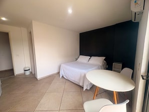 A hotel room with a bed, a small table, a chair, a trash bin, and a wall-mounted air conditioning unit. - Stratus Centro Hotel (Volta Redonda)