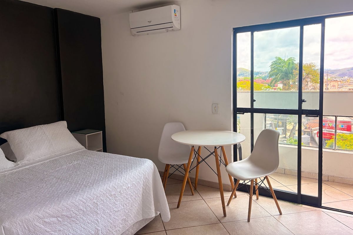 A bedroom with a bed, a small table, two chairs, a balcony with a view, and an air conditioning unit.