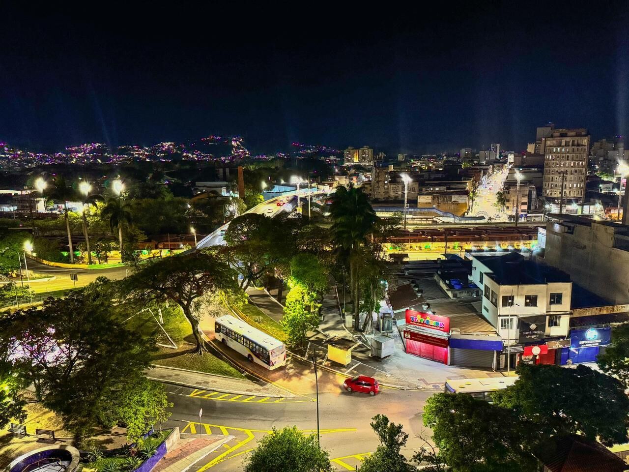 Un paisaje urbano nocturno con una estación de autobuses, una estatua y un edificio con un letrero rojo.