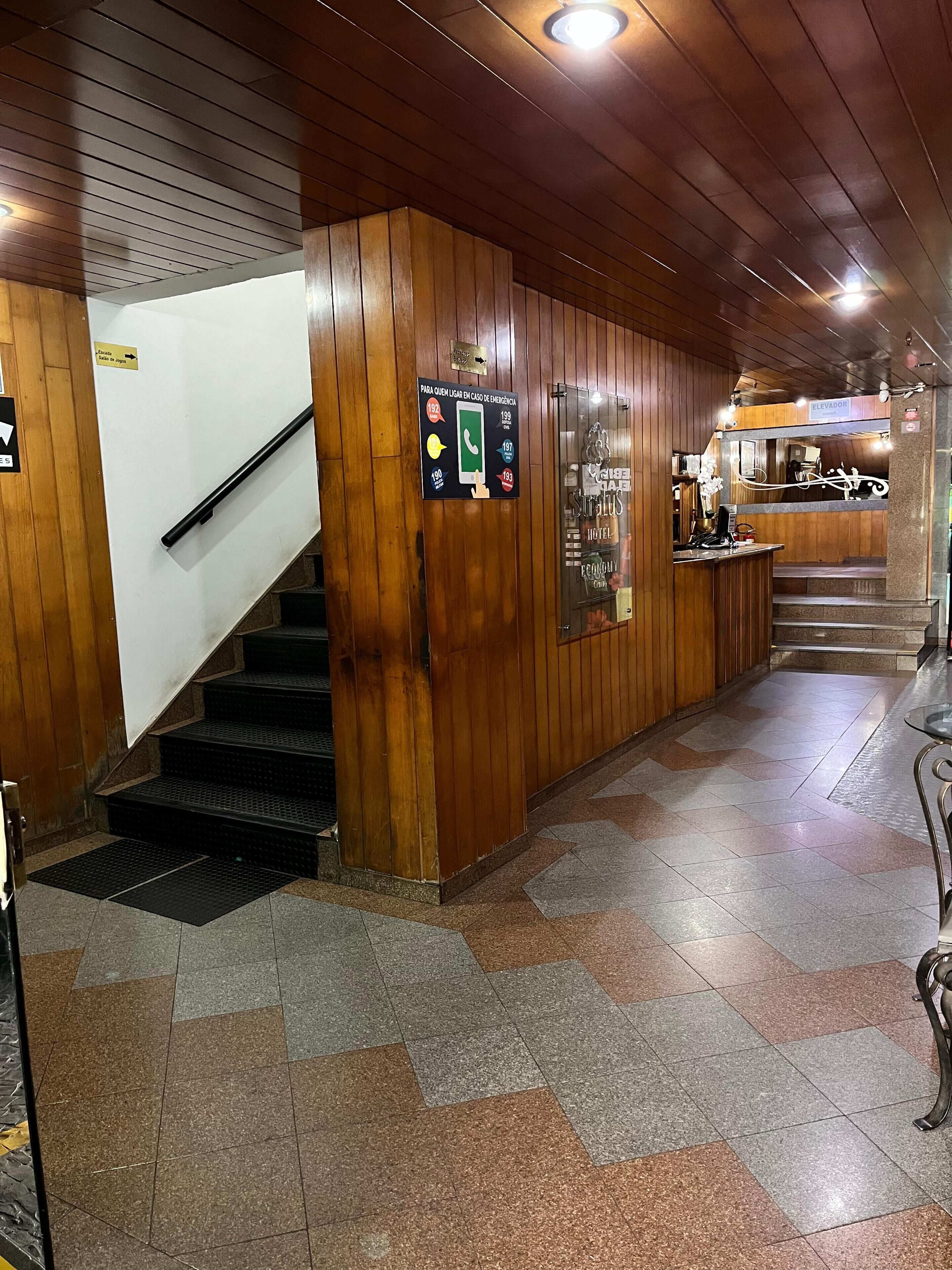 Un vestíbulo de hotel con paneles de madera, una escalera, una recepción y señales de contacto de emergencia.