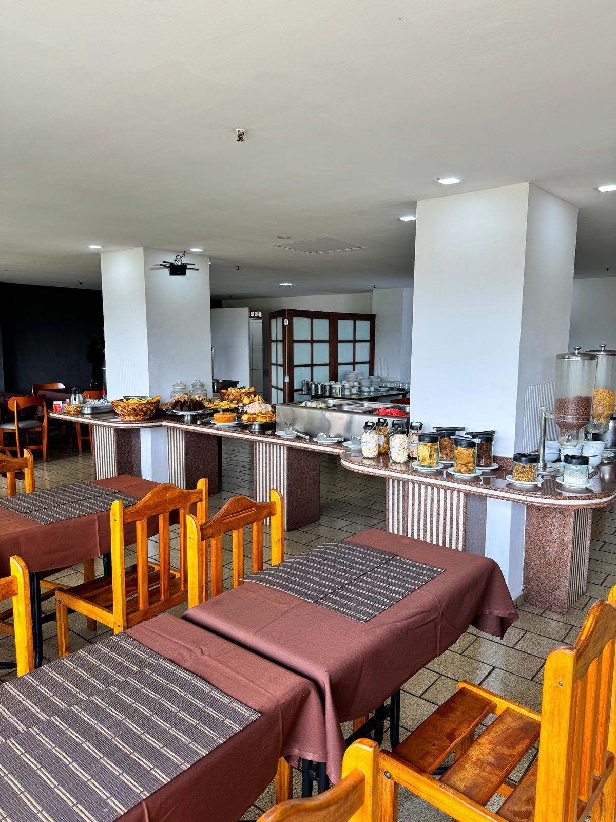 Un restaurante con varias mesas y sillas, un mostrador de buffet con diversos alimentos y una zona de cocina al fondo.