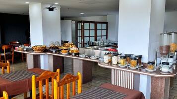Un restaurante con varias mesas y sillas, un mostrador de buffet con diversos alimentos y una zona de cocina al fondo.