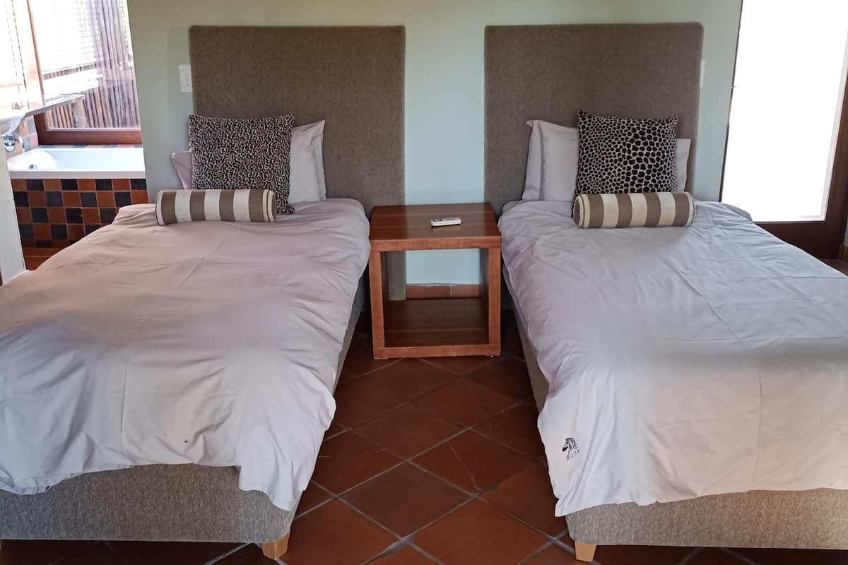 Ein Zimmer mit zwei Betten, jeweils mit Kopfteil, einem kleinen Tisch und Blick auf einen Balkon.