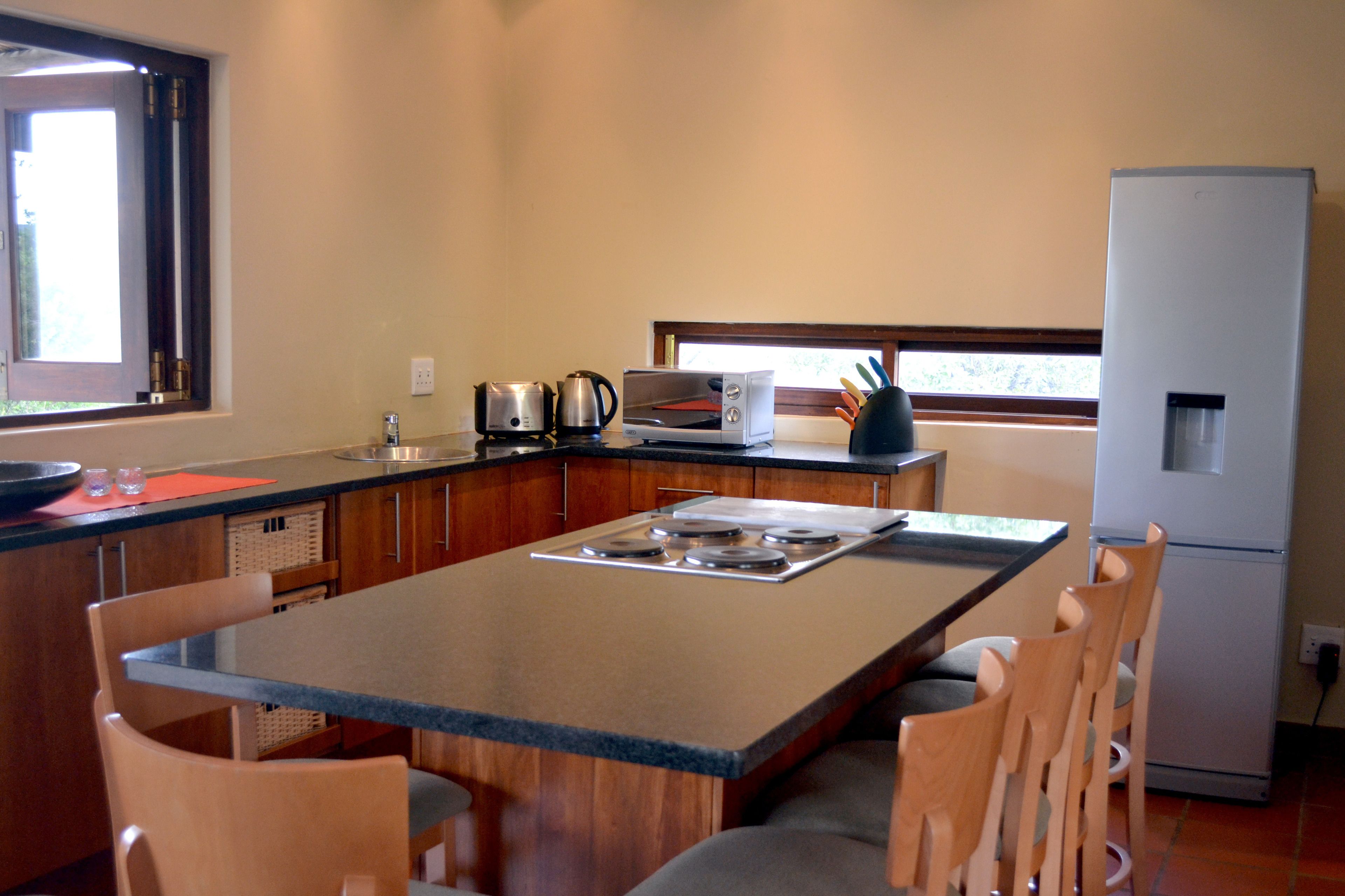 Cocina compacta con una isla central, sillas de madera, horno integrado, refrigerador y ventana que proporciona luz natural.