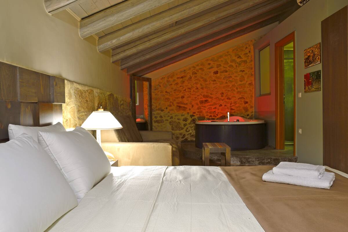Un dormitorio con una cama, una mesita de noche, una lámpara, una silla, una pequeña mesa y una pared de piedra.