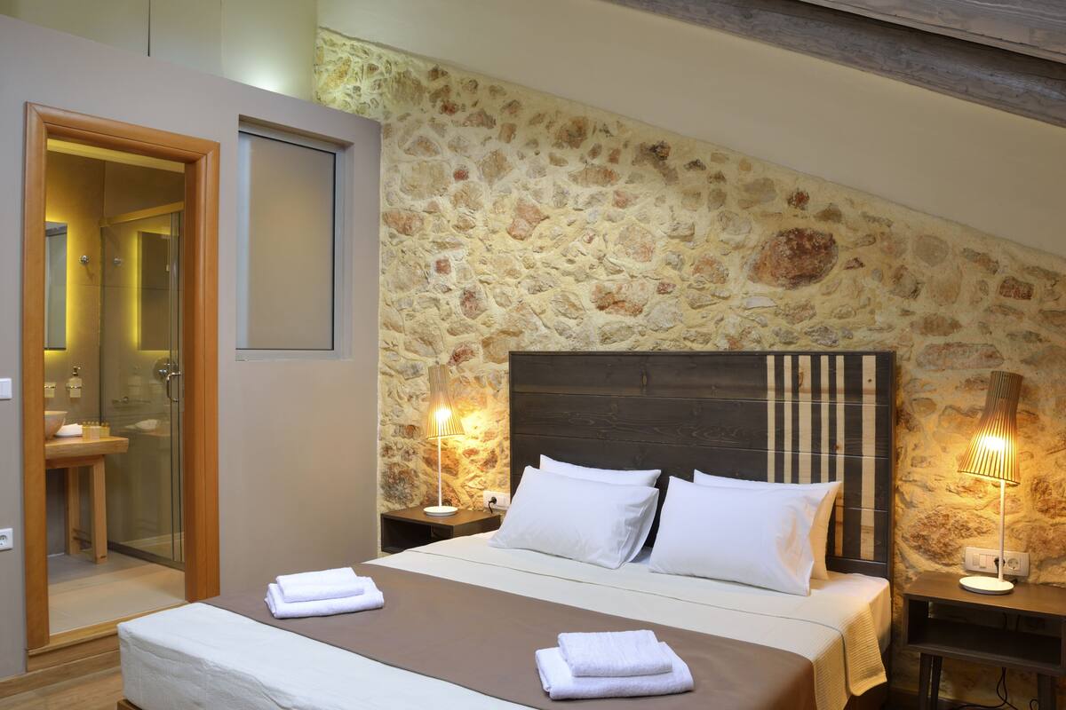 Un dormitorio con pared de piedra, cabecera de madera, cama con ropa blanca y una mesita de noche con lámpara.