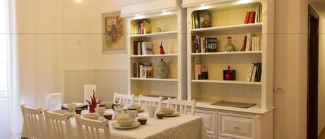 A dining area with a table set for four, surrounded by white chairs. There are bookshelves with various books and decorative items.