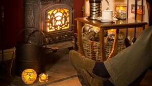 A cozy room with a wood-burning stove, a table with a cup and saucer, and a basket filled with items. - Hermiston Guest House (Keswick)