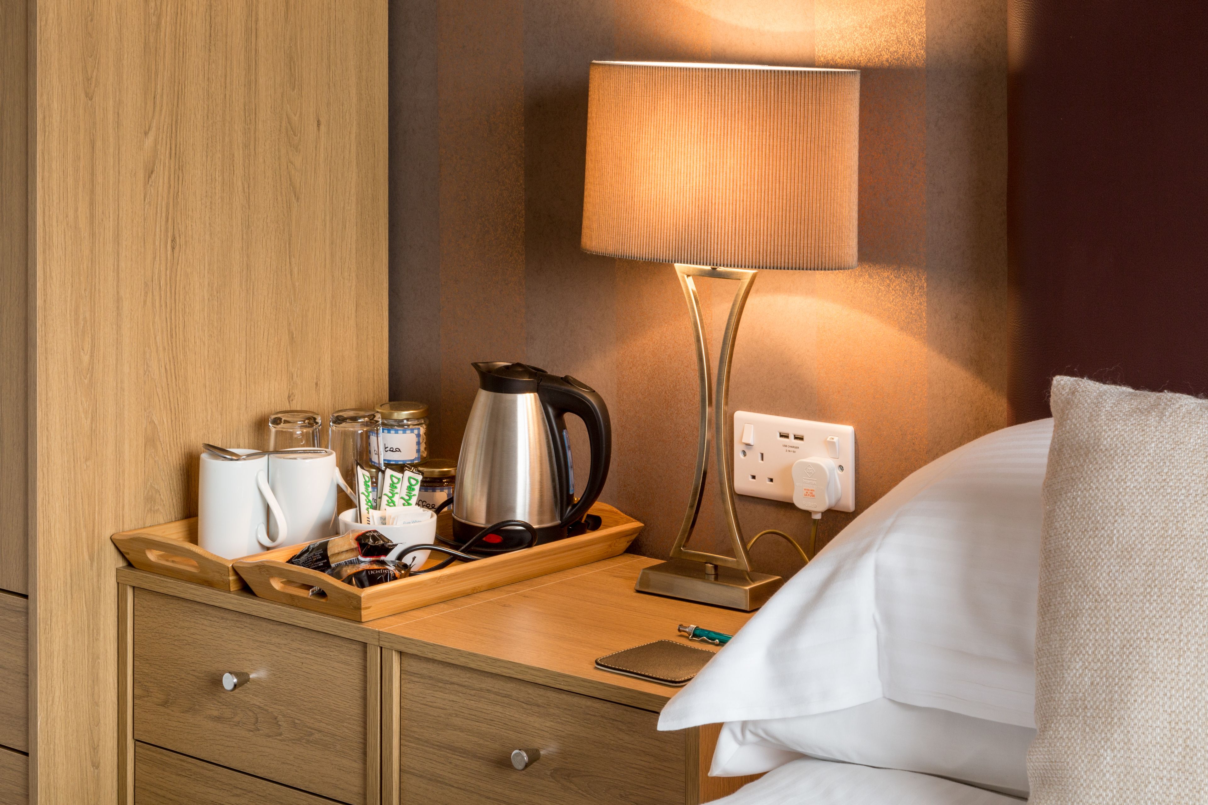 A hotel room with a wooden nightstand, a lamp, a kettle, a tray with snacks, and a bed with white linens.