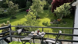 A balcony with a table set for two, overlooking a well-manicured garden with various shrubs and a lawn. - Hermiston Guest House (Keswick)
