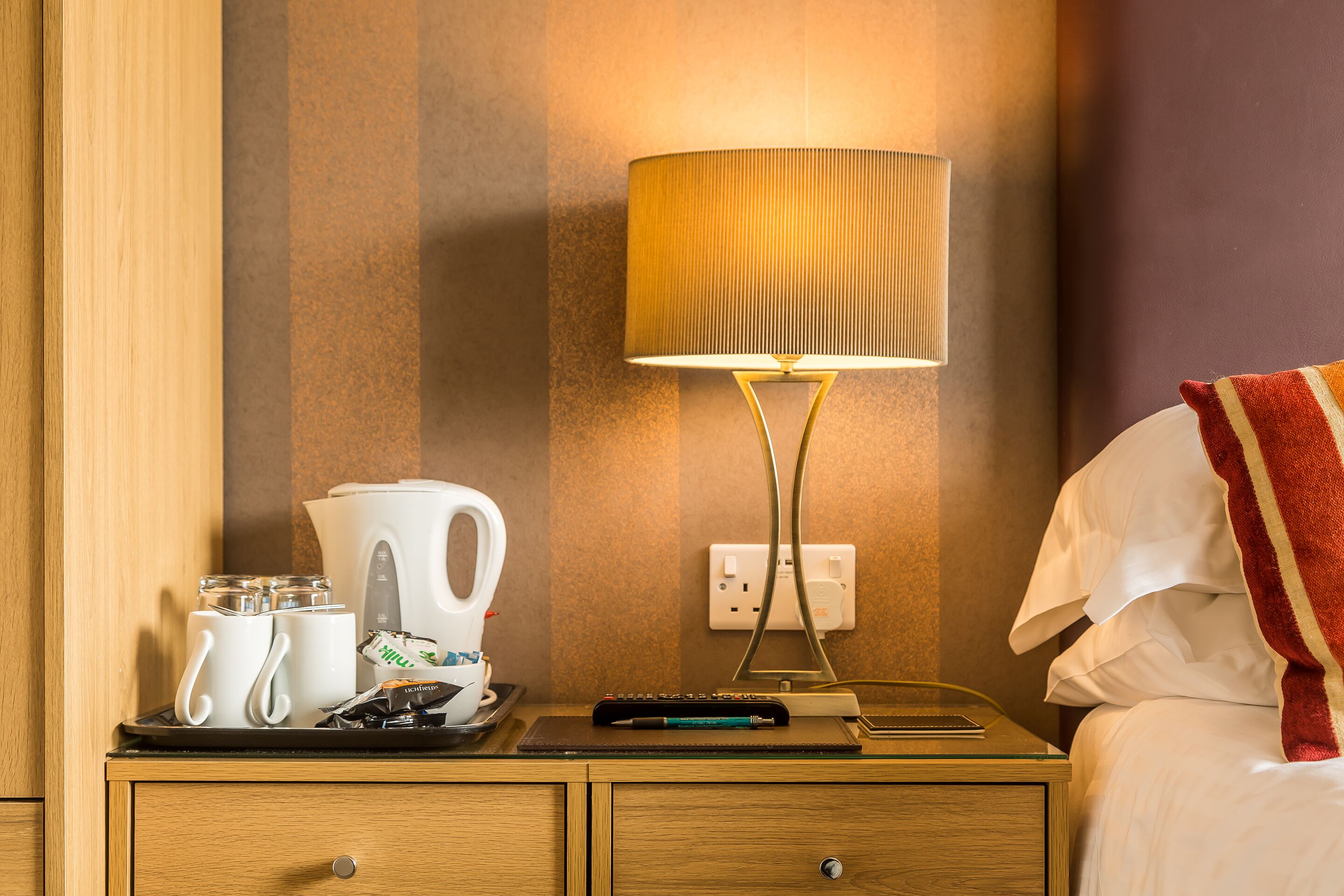 A hotel room with a wooden nightstand, a lamp, a tray with mugs, a coffee maker, and a remote control.