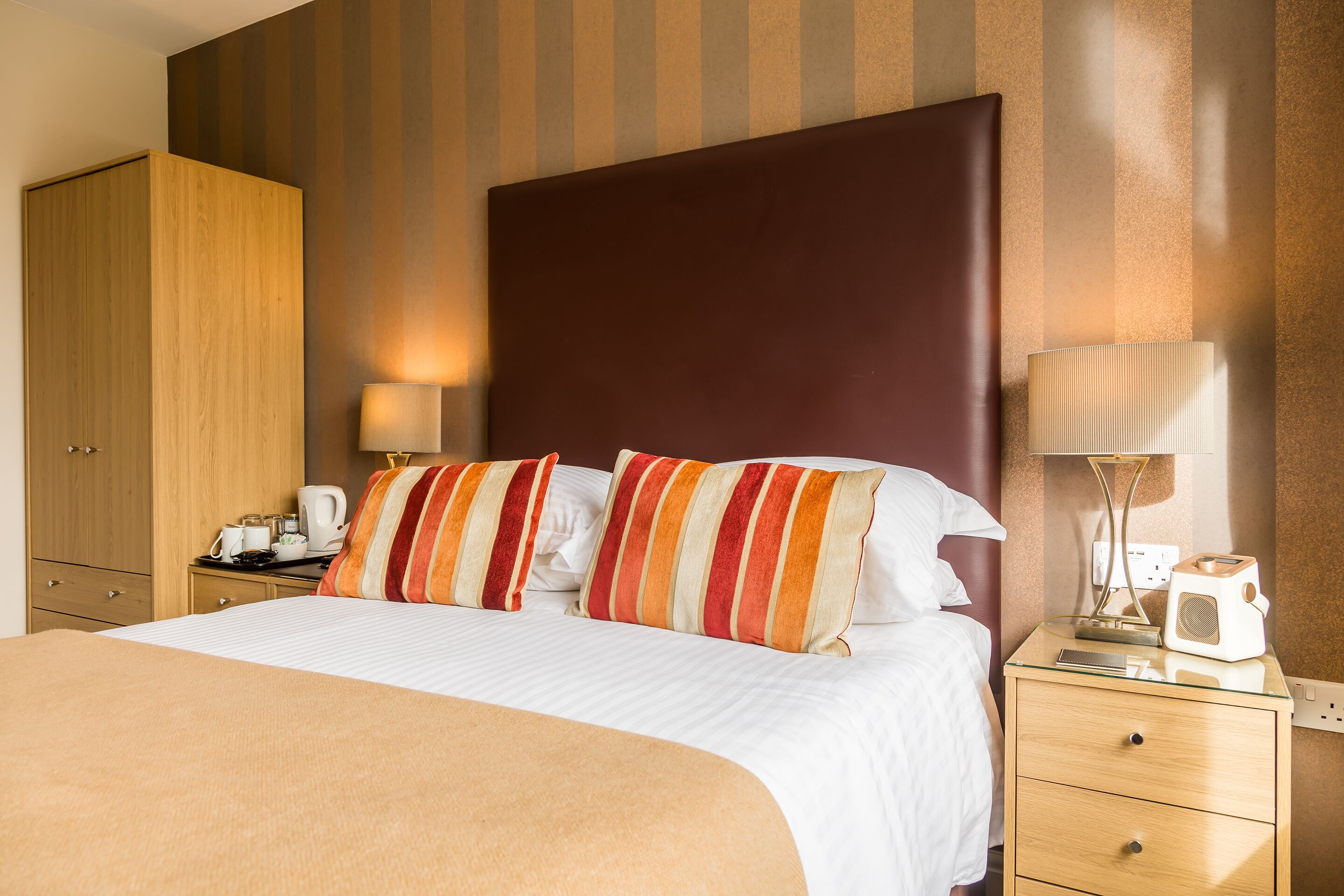Una habitación de hotel con una cama grande, almohadas a rayas, un armario de madera, mesitas de noche con lámparas y una mesita auxiliar con una radio.