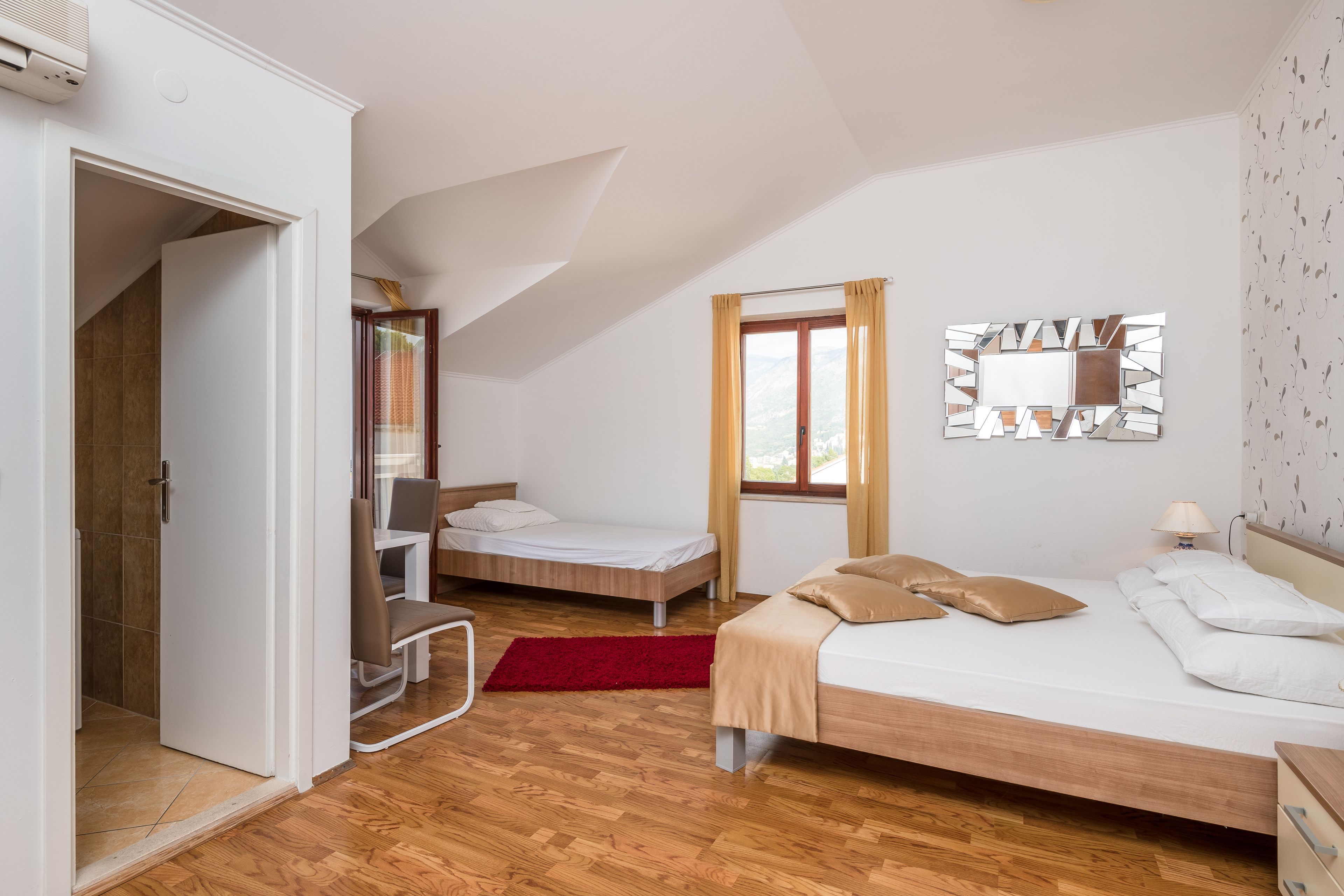 A bedroom with a wooden floor, a bed with beige bedding, a red rug, a window with curtains, and a mirror on the wall.