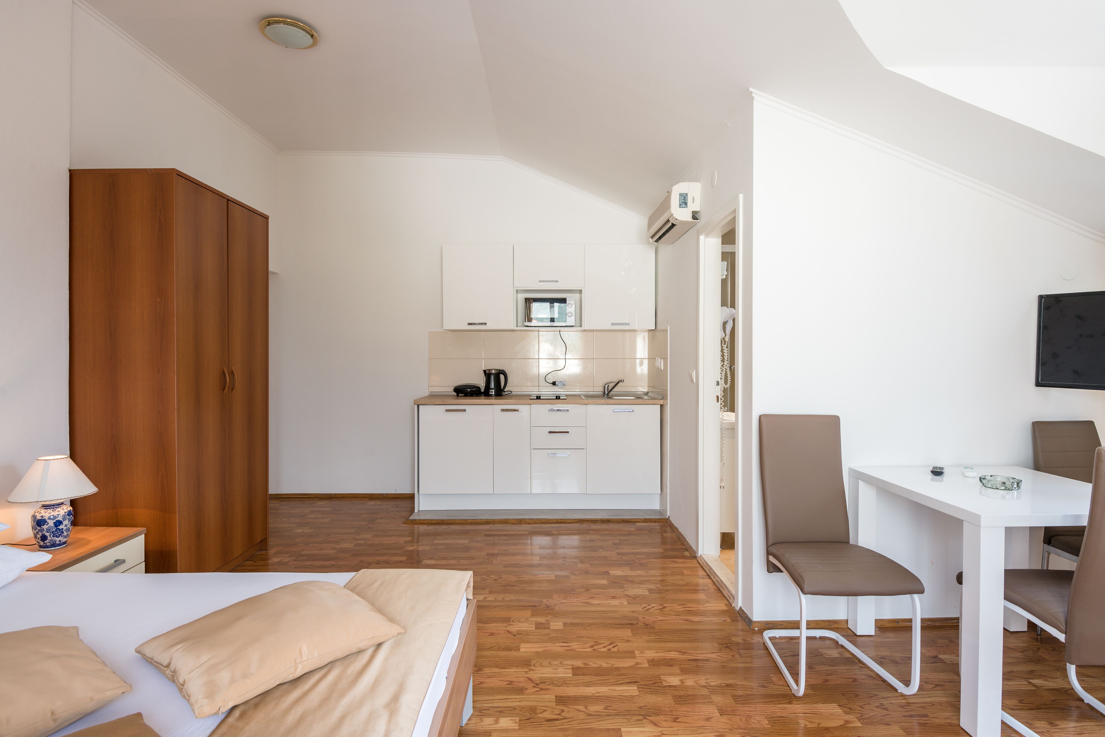 A compact hotel room with a bed, a desk, a chair, a kitchenette, and a wardrobe.