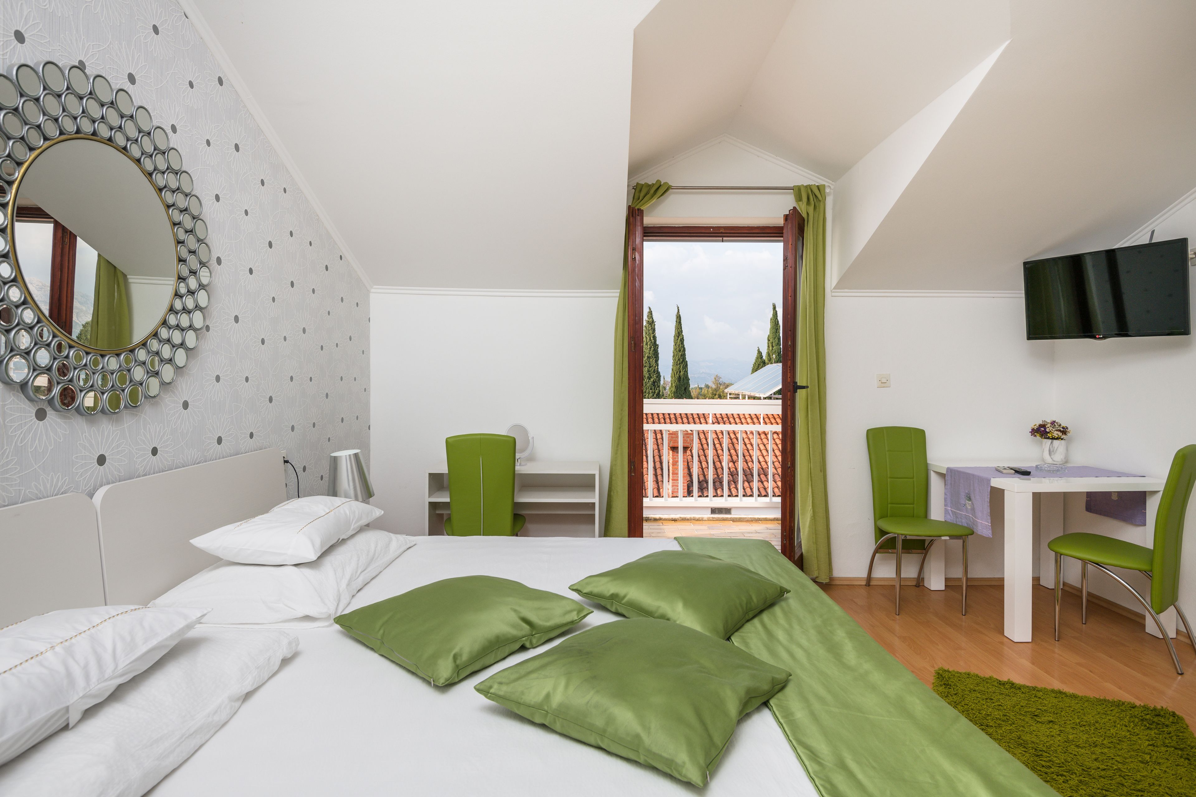 A bedroom with a bed, green pillows, a desk, chairs, a TV, and a balcony with a view.
