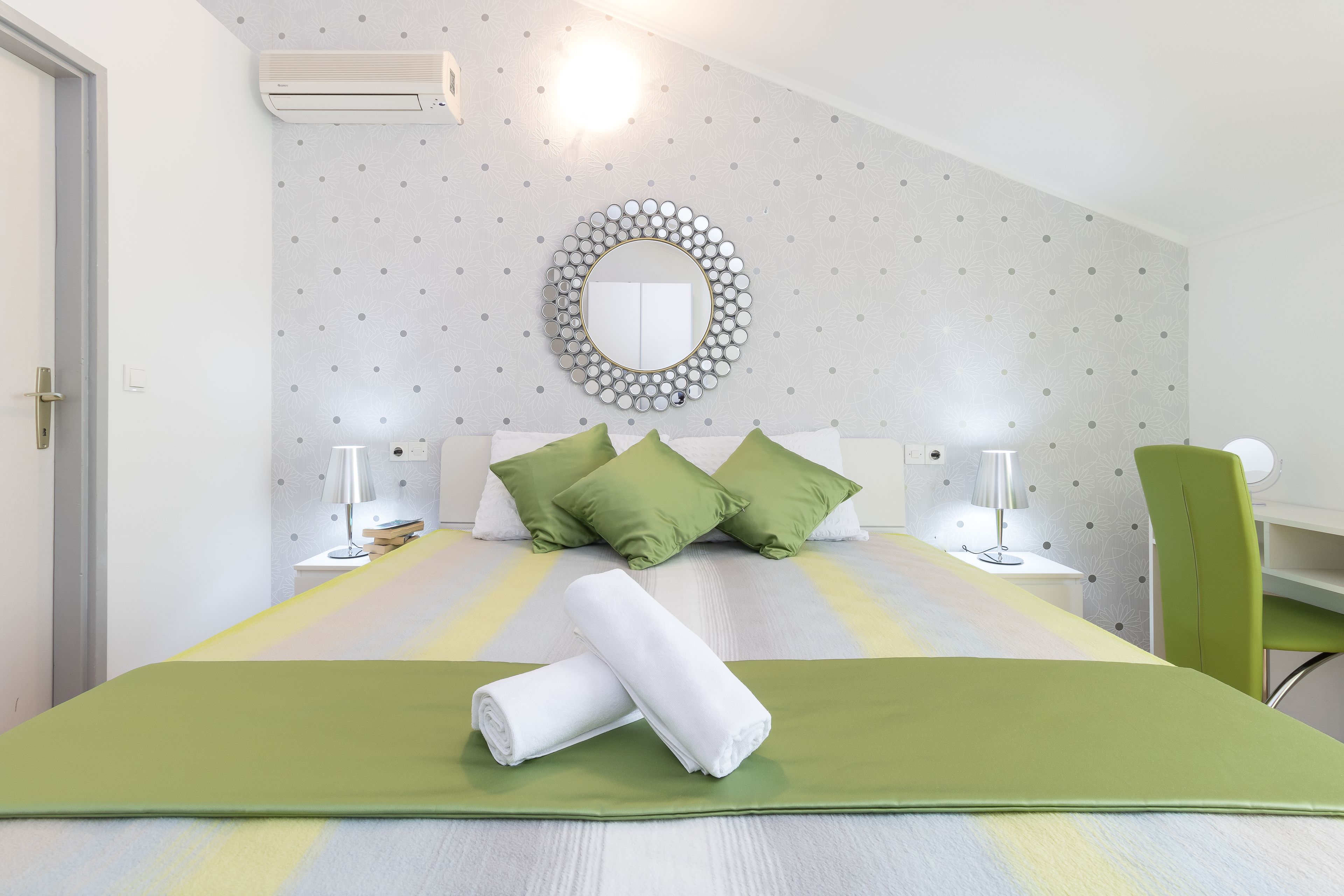 A neatly made bed with green and white bedding, a round mirror on the wall, and a desk with a chair.