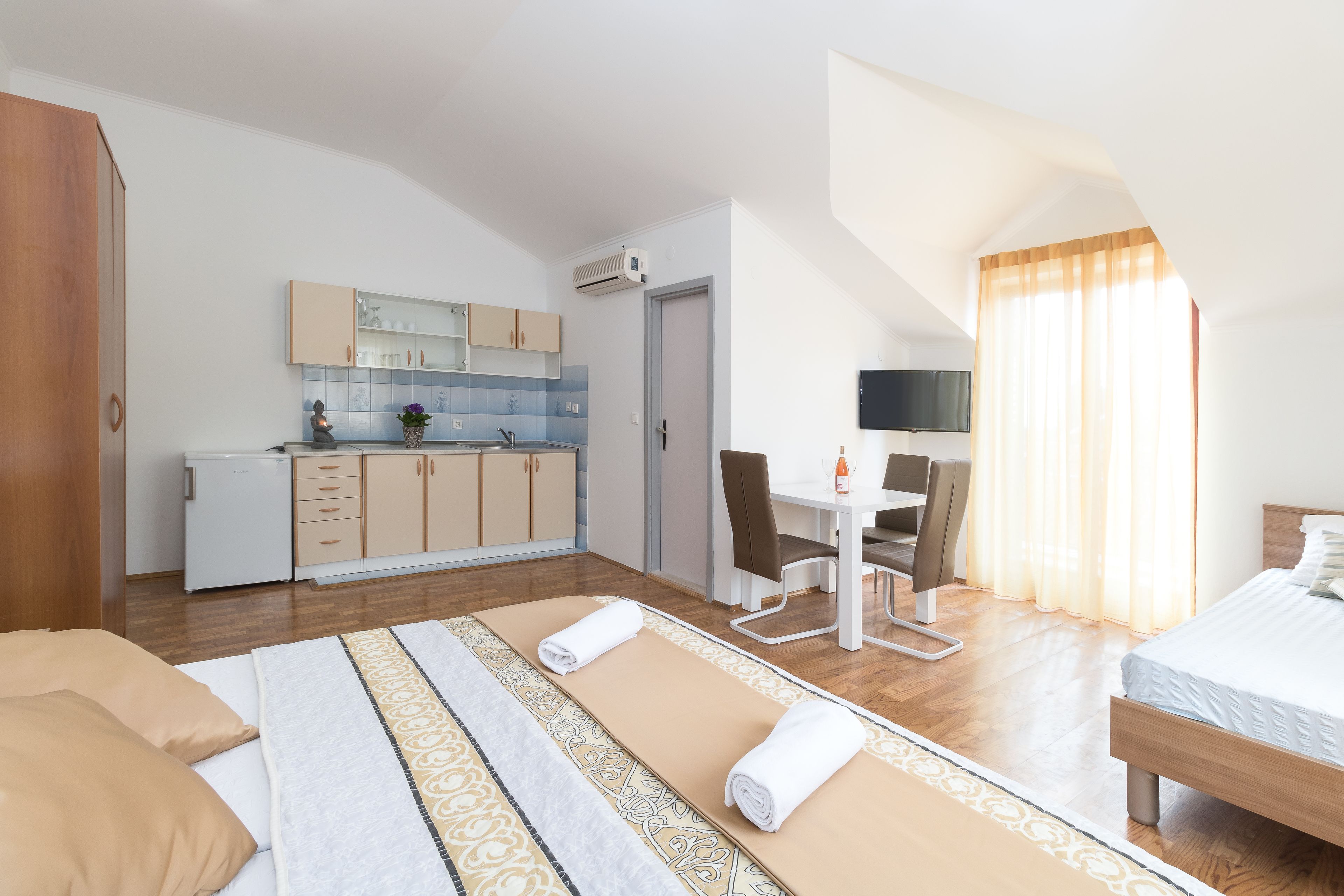 A compact hotel room with a bed, a desk, a chair, a small kitchenette, and a TV.