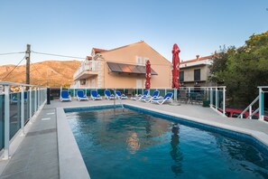 A swimming pool with blue lounge chairs, a building with a balcony, and a red umbrella. - Villa Voinić (Konavle)
