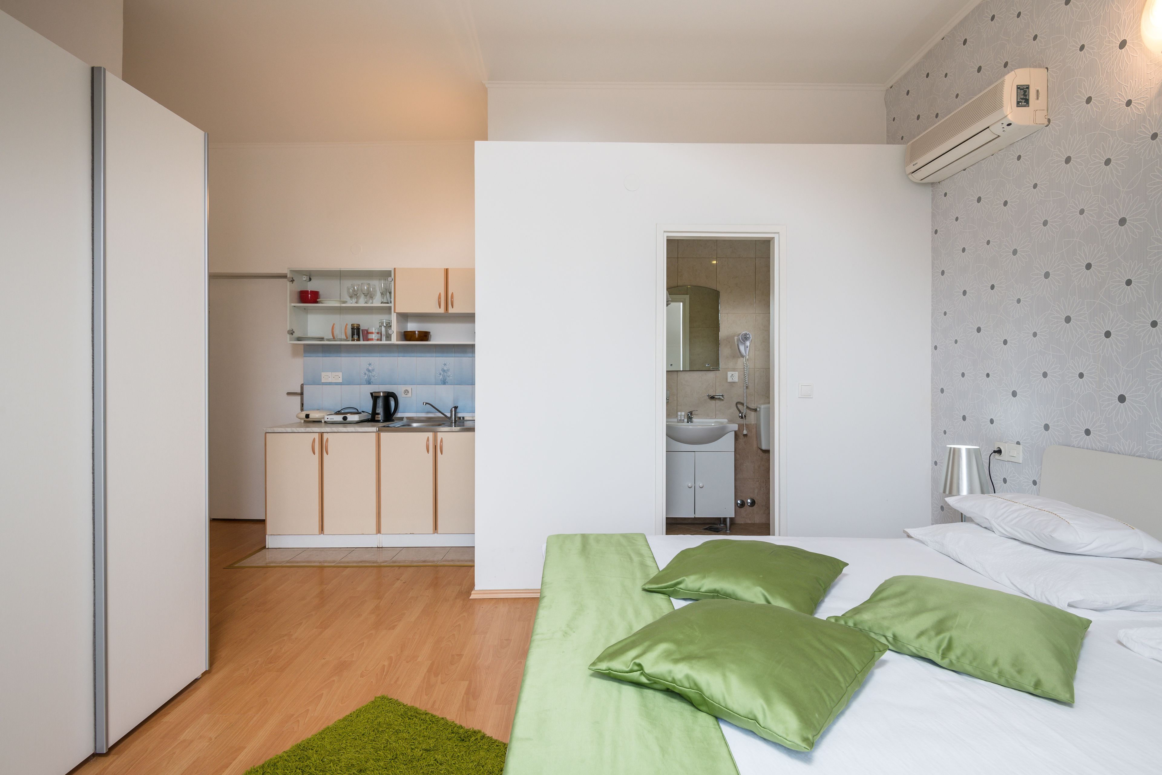 A compact hotel room with a kitchenette, a bed with green pillows, and a bathroom visible through an open door.